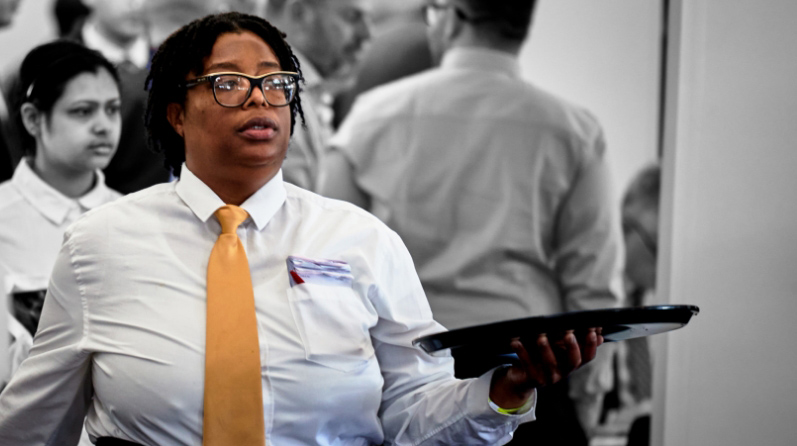 A Black woman balances an empty drinks tray on an upturned hand. She is wearing a white shirt, black-rimmed glasses and yellow tie.
