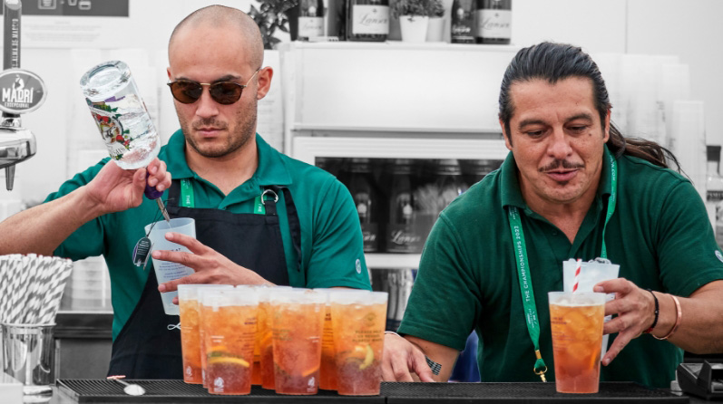 Two men, wearing dark green polo shirts, standing side by side behind a bar; they are making cocktails.