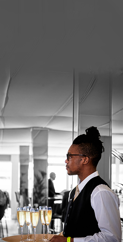The side profile of a young Black man holding a tray of champagne flutes filled with sparkling white wine. He has dreadlocks, is wearing glasses, a white shirt and a black waistcoat.