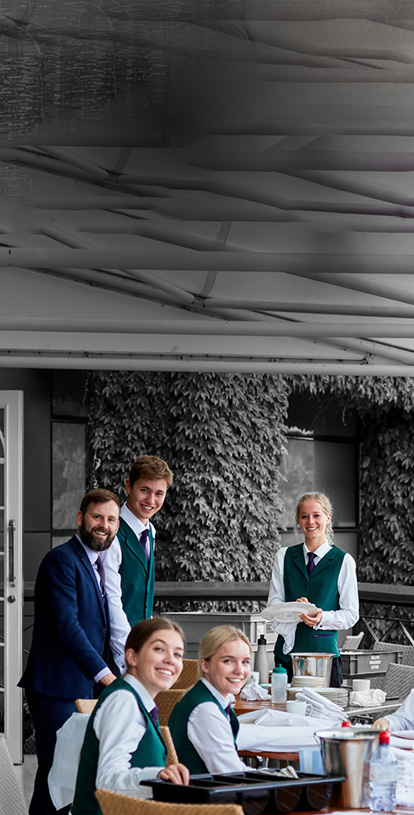 A collection of people in smart uniforms are stood or sat around some tables, facing the camera and smiling. A White man is stood wearing a navy blue suit. The rest are slightly younger, wearing white shirts with green waistcoats and purple ties.