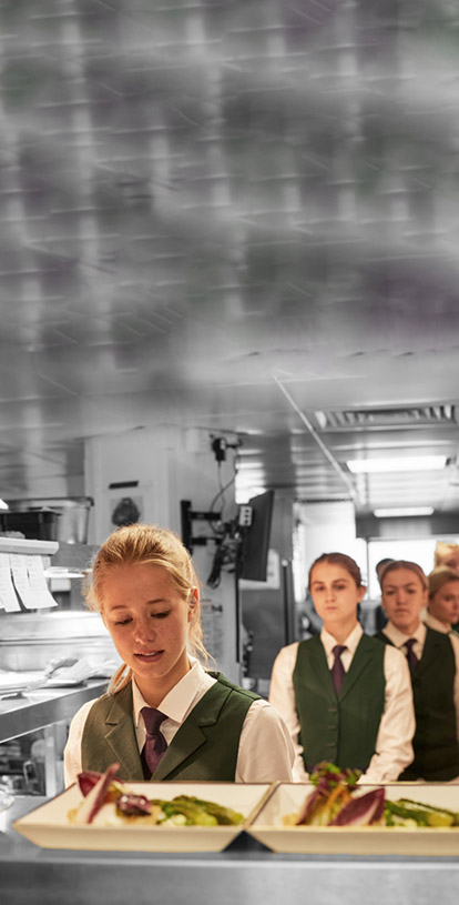 A young White woman with blonde hair, wearing smart wait staff uniform, is about to pick up some well presented, plated food. Behind her is a queue of young women in the same attire awaiting the same.