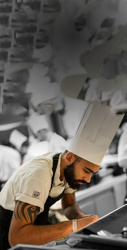 A man in chef's uniform is bent over a worktop preparing food. He has a full, dark beard, and is concentrating on his task.