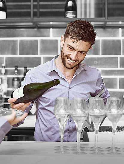 In focus is a smiling White man with brown hair. He wearing a purple shirt and is pouring sparkling wine into some champagne flutes. In the foreground a blonde female is facing towards him, holding a drinks tray